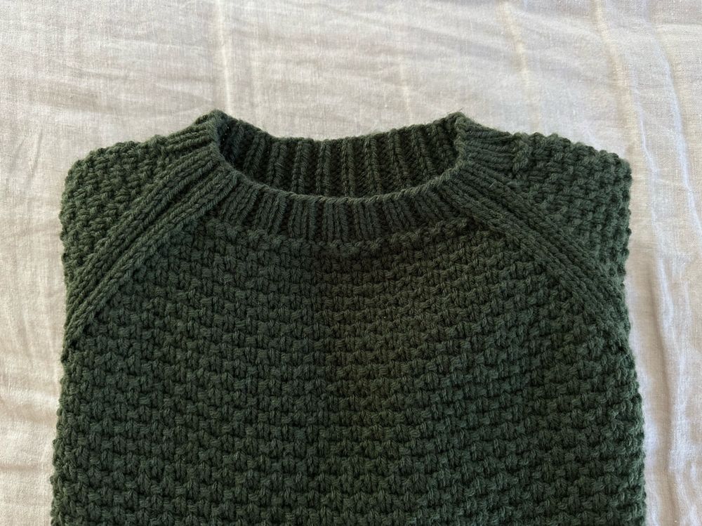 A green, textured hand knit sweater that has been folded and placed on a linen duvet. 