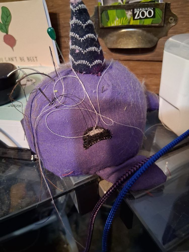 Purple Narwhal plush with a black horn and pins and needles sticking out of it. 