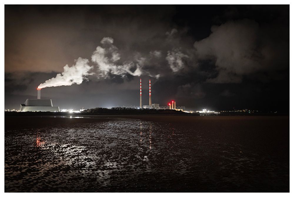 A night time photo, taken on Sandymount beach, looking towards the Poolbeg towers. To the left is the Dublin waste incinerator, sending plumbs of smoke from its chimneys off to the right and over the towers, which are located in the center of the image. To the right, the lights of Clontarf and Howth are visible, off in the distance across Dublin bay. The tide is out, and little pools of water between the rippling sand reflect the scene above