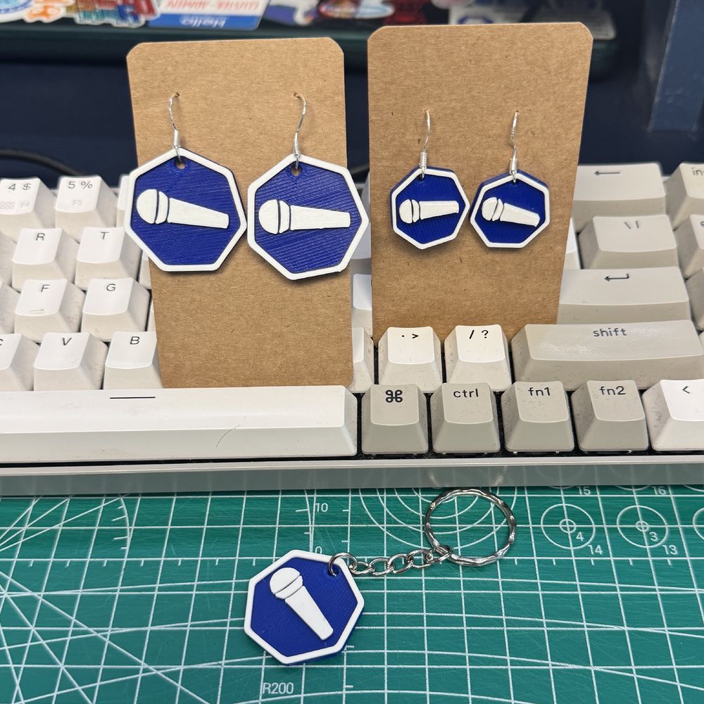 A photo of some 3D printed earrings in the shape of the Kuberoke logo hanging in some small card leaning against a keyboard. There are two sizes of earrings show. And below both a keyring featuring the same logo. 