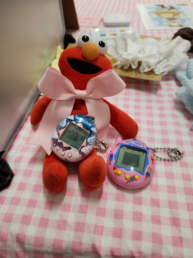 A small plush elmo sitting on my table at an event. There's a tamagotchi angel on his lap and a connection next to him.