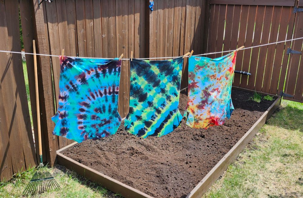 Three tie-dyed t-shirts hanging from a clothesline. In the background is a fence and an empty garden plot. 