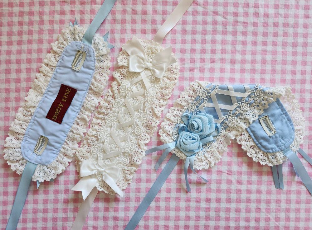 Three rectangle headdresses from bodyline on a pink gingham background. From the left there are two smaller headdress in blue and white, one is flipped upside-down showing the sewn-on weft clips, the second is right side up to show the sewn down ribbons. On the right is a larger blue ribbon-rose headdress, it's folded diagonally to show both the decorative ribbons and the sewn clips on the back.