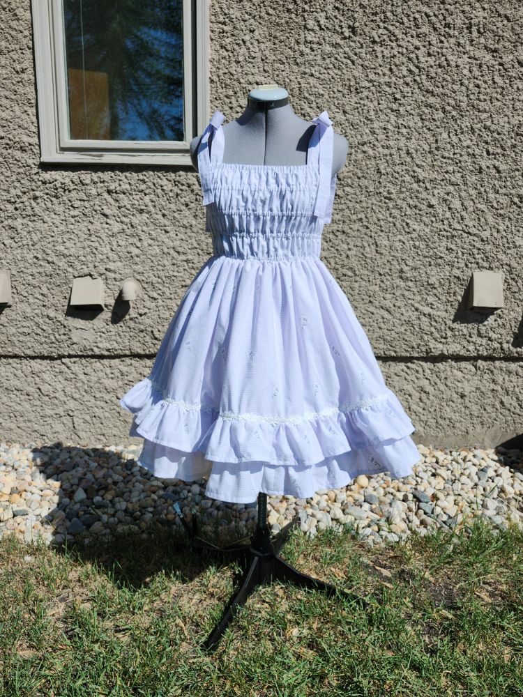 A light purple jsk photographed outside in front of a wall. It has a fully shirred bodice and the straps tie in bows at the shoulders. The skirt has two ruffles at the bottom with a white floral trim at the top of the first ruffle.