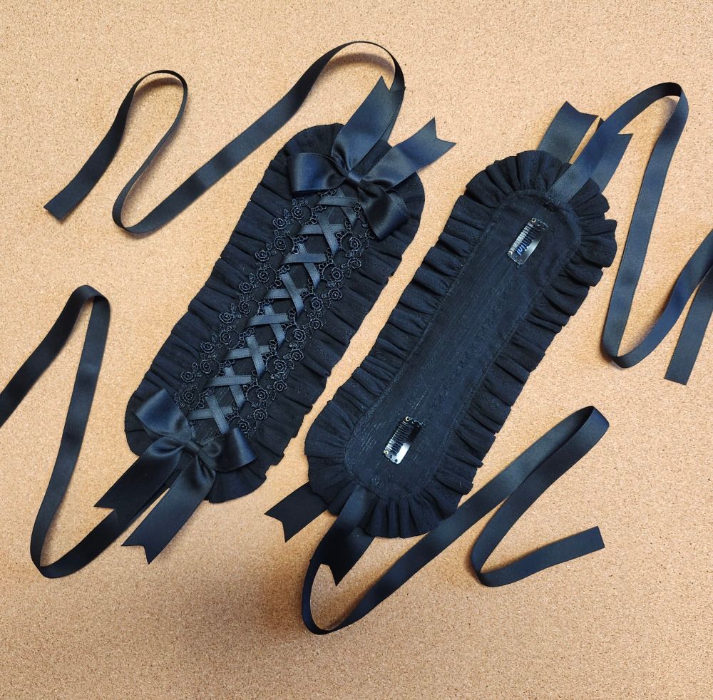 Two black flat/rectangle style egl headdresses. One is right side up to show details (criss-cross ribbon lacing from a rose trim, and a larger bow at each end) and the other is flipped over to show the clips sewn to the back. The ruffle around the outside is made from the same fabric and the backing. Both have the ribbon ties filling the sides of the image in a squiggle.