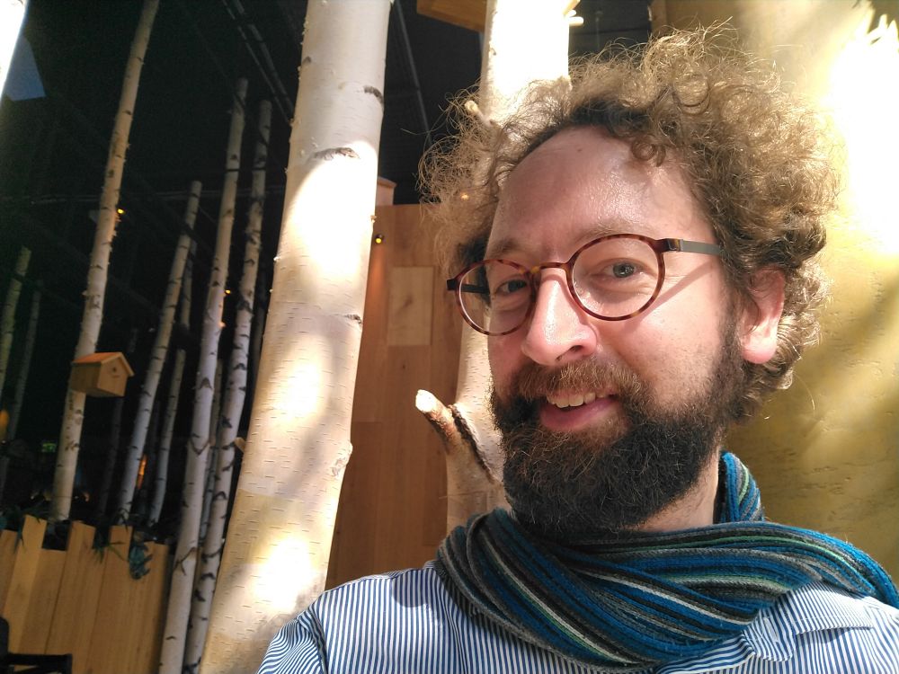 Manuel lächelt in die Kamera. Hinter ihm Birkenstämme. An einem davon hängt ein Vogelhäuschen.