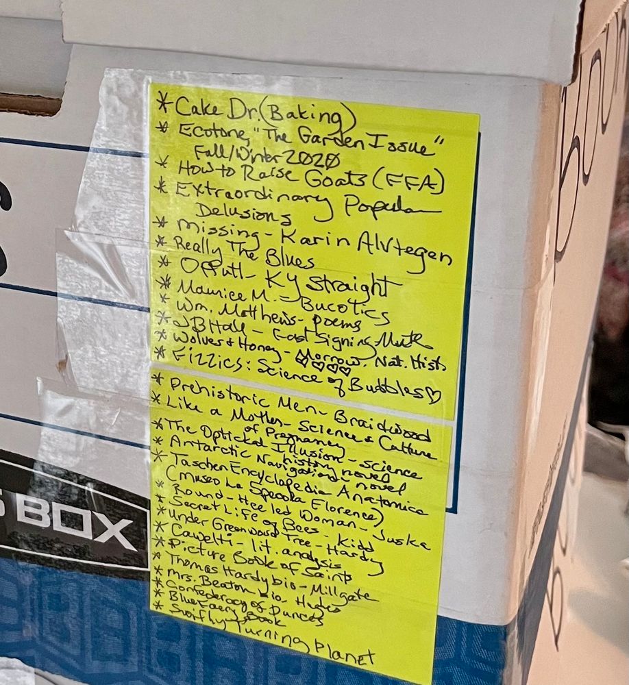 Close-up of a bankers box with 2 post-it notes taped to the side. Each neon yellow post-it contains 12-15 book titles scribbled by hand, indicating the contents of the box.