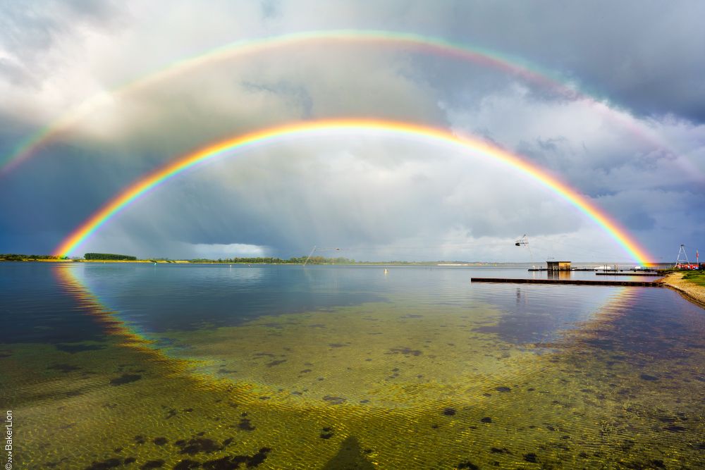 Doppelter Regenbogen von einem zum anderen Ufer am Veerse Meer Holland vor bewölktem Himmel mit drittem spiegelnd im glatten Wasser 