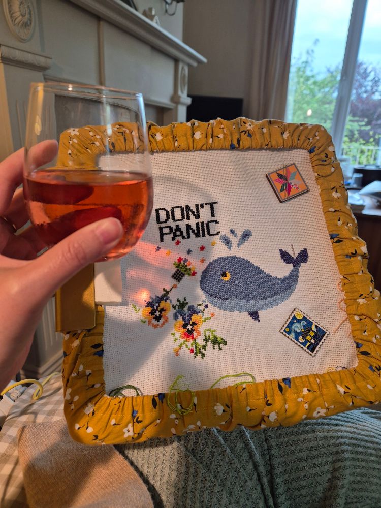 Left hand holding a tumbler of pink liquid. Cross stitch in the background showing a whale and petunias with the words Don't Panic. 