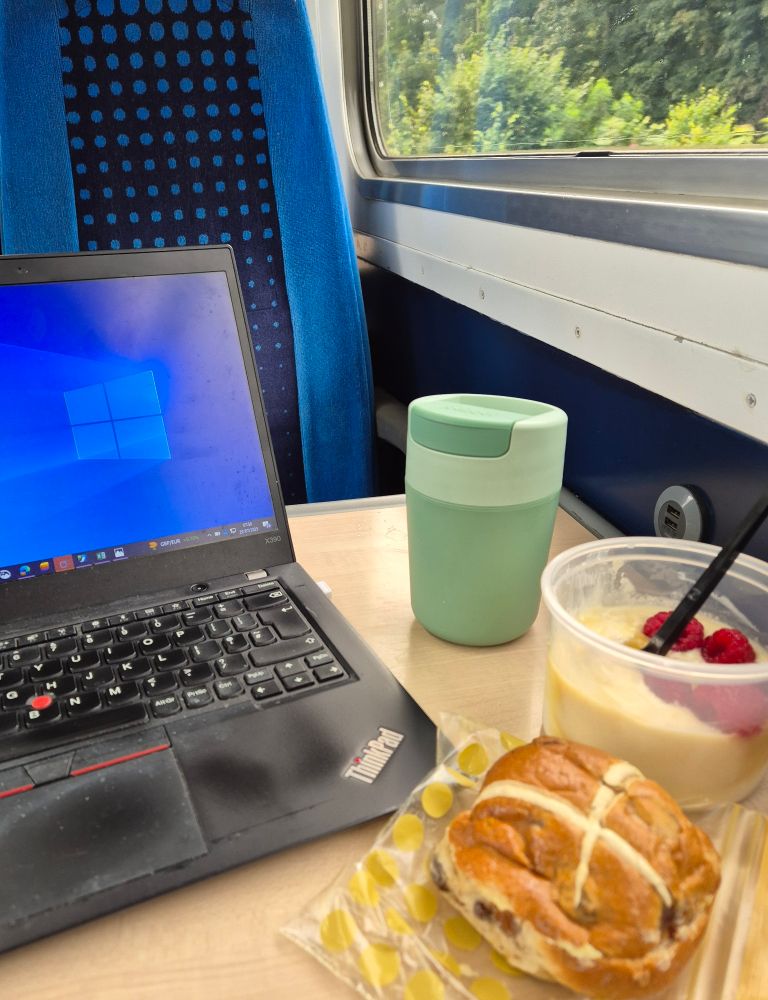 Showing a table on a train. On the left is a laptop. On the right a travel coffee mug, a pot of yoghurt and raspberries and a hot cross bun. 