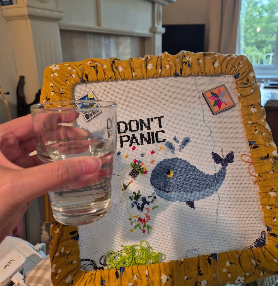 Left hand holding a tumbler of clear liquid. In the background is a partially completed cross stitch showing a whale, a bowl of petunias and the words 'Don't Panic'. 