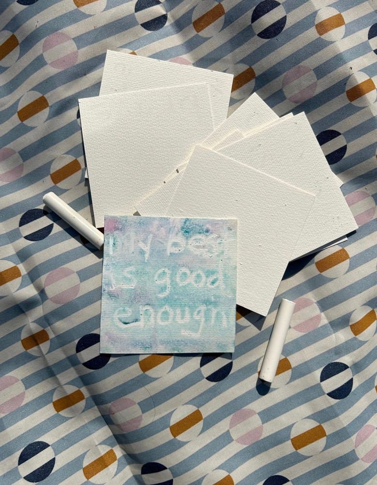 A geometric background with a pile white square cards, white crayons, and a card on top painted with watercolor. In white is a handwritten message: My best is good enough. 