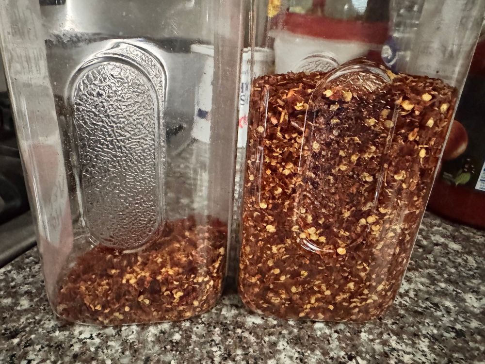 A picture showing the same two containers from the side, so you can see the contents. If you don’t know the difference, they look almost identical in color and texture. The left container is only about 10% filled (I need to crush more dried Thai chilies), and the one on the right is about 75% full of crushed red pepper. Both are a vibrant red pepper flake, with seeds visible.