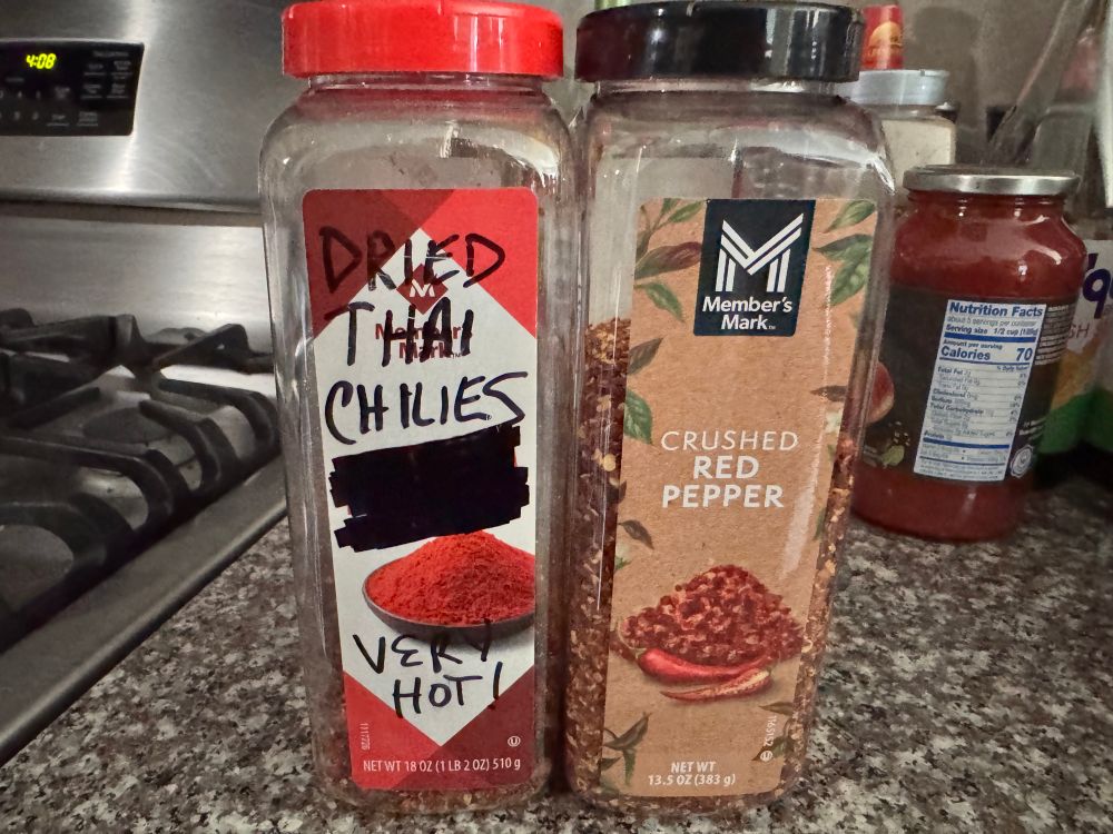 A picture showing two large containers of crushed red chilies. The one of the left is labeled with black marker as “Dried Thai Chilies - Very Hot!” And the one of the right is simply “Crushed Red Pepper.”