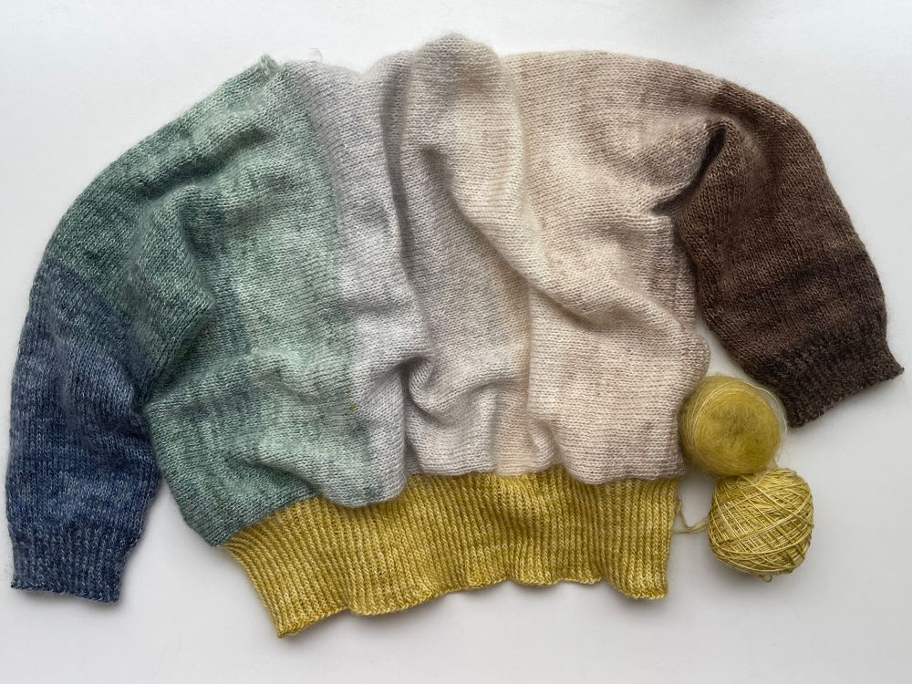 Knitting and yarn against a white background

The body of the cardigan is displayed with its back and sleeves visible. The left sleeve cuff is a dark blue and this colour progresses along the sleeve to dark greens then across the body it shifts to lighter greens, greys, creams and soft caramel tones before the right sleeve where the brown tones deepen to a dark brown cuff.

The hem is a bright golden mustard colour (i find it hard to describe!) the matching balls of yarn are at the bottom right of the photo. They will be used for the button band