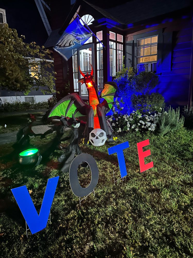 Halloween display of three witches around a glowing green cauldron with a dragon behind them. A VOTE sign is in front of it. 