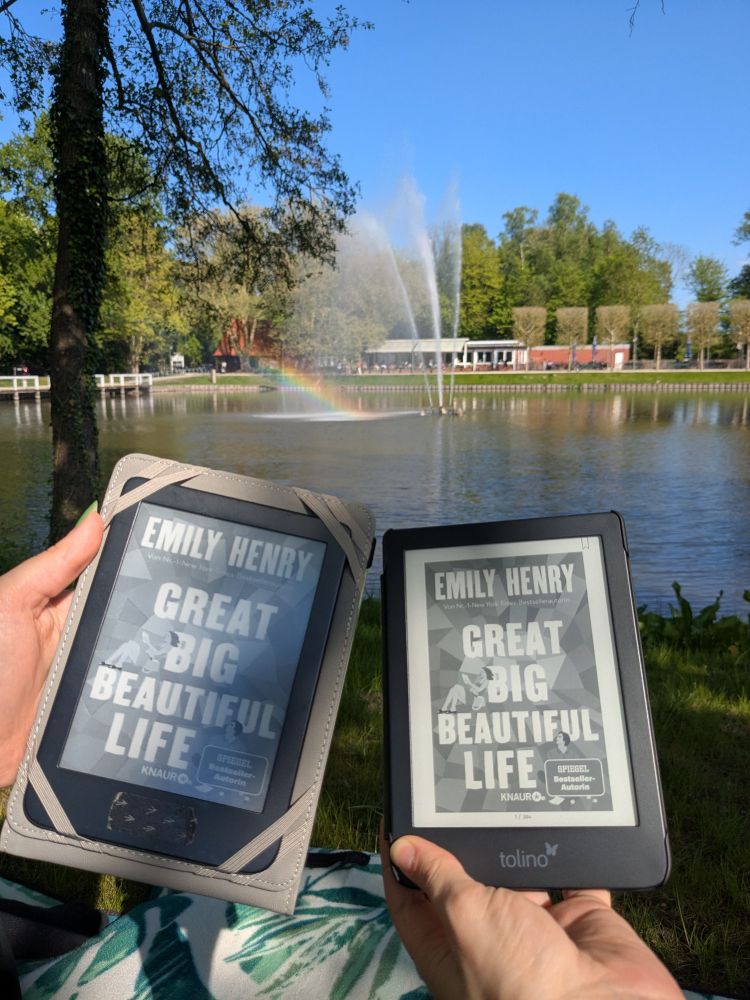 Im Vordergrund zwei e-reader mit dem Buchcover von "Great Big Beautiful Lie" von Emily Henry. Im Hintergrund bisschen grün und eine Wasserfontäne, die einen kleinen Regenbogen produziert