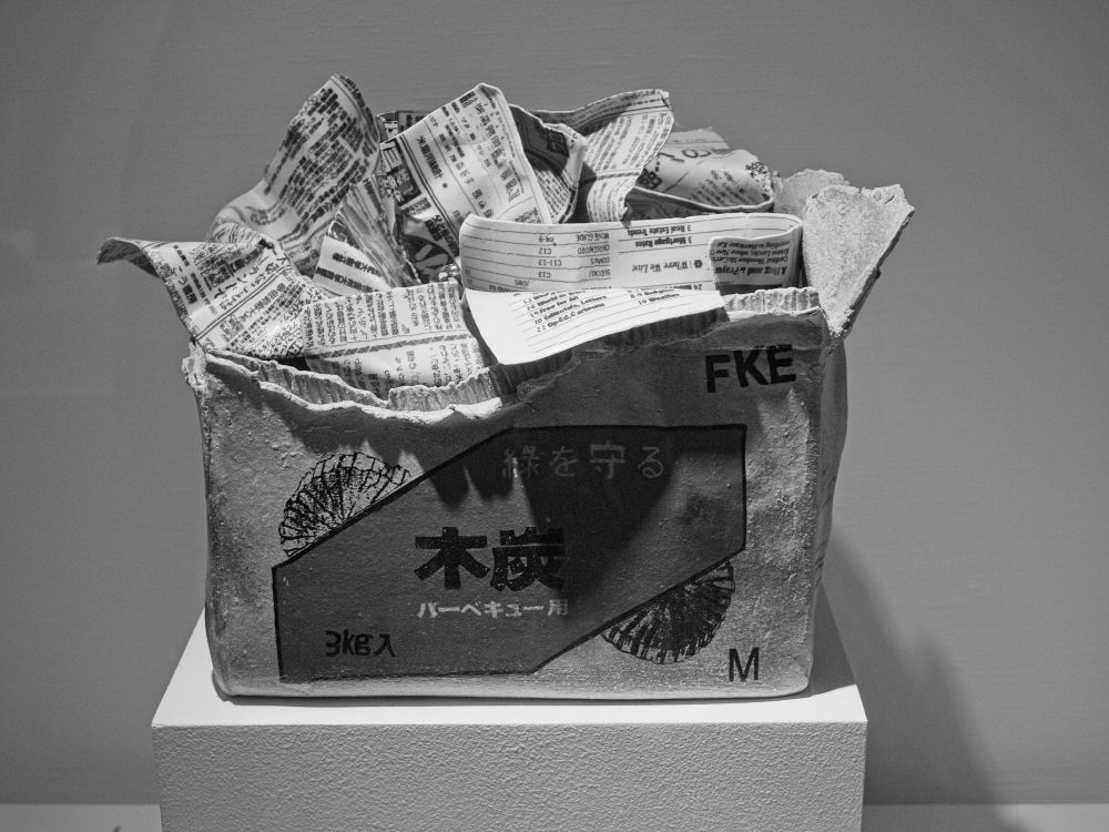 Black and white image of a sculpture of a box with newspapers protruding from it.