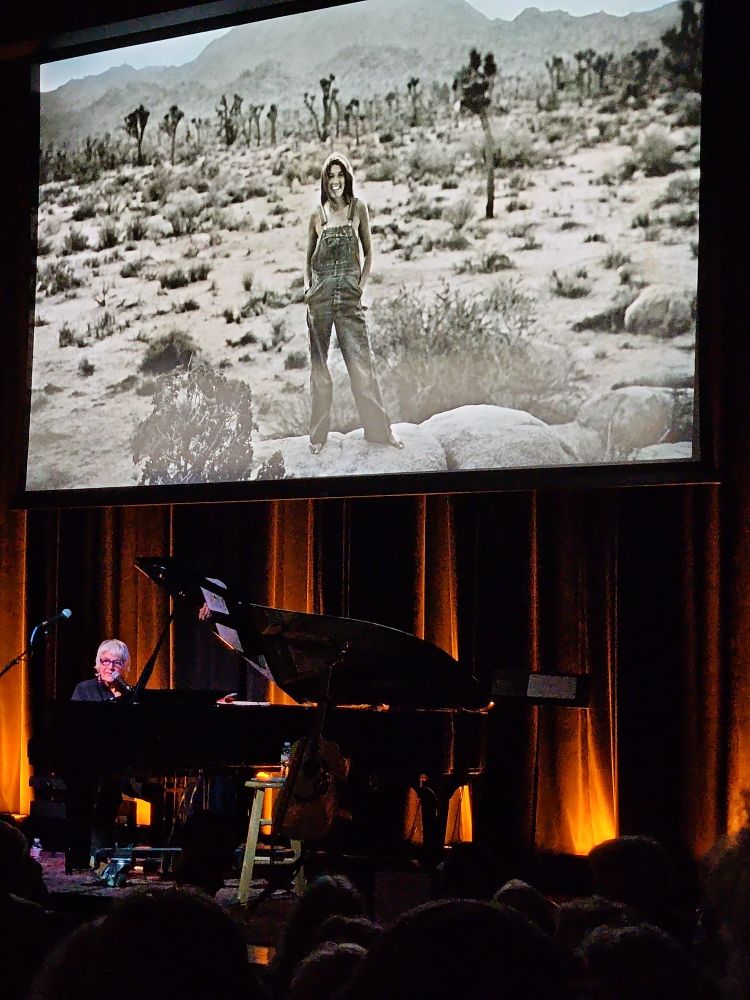 A black & white picture of a young woman standing in the desert in overalls smiling, hangs above the same woman at her piano, now 50 years older and stll smiling.
