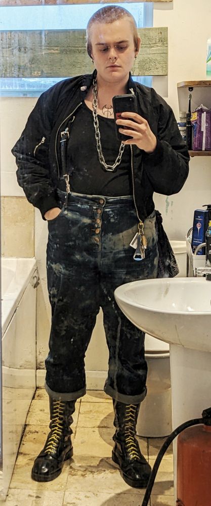 leatherdyke with #2 buzzcut wearing a bomber jacket, tank top, leather braces, heavy chain, bleached jeans, and 20-eye solovairs with yellow laces