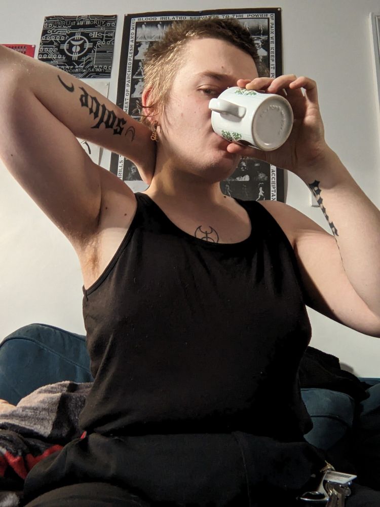 butch trans woman with boyish scrungly bed hair, gold facial piercings. wearing a black tank top which exposes a labrys chest tattoo. drinking from a cup of coffee with hand placed on the back of her neck, elbow outstretched.
