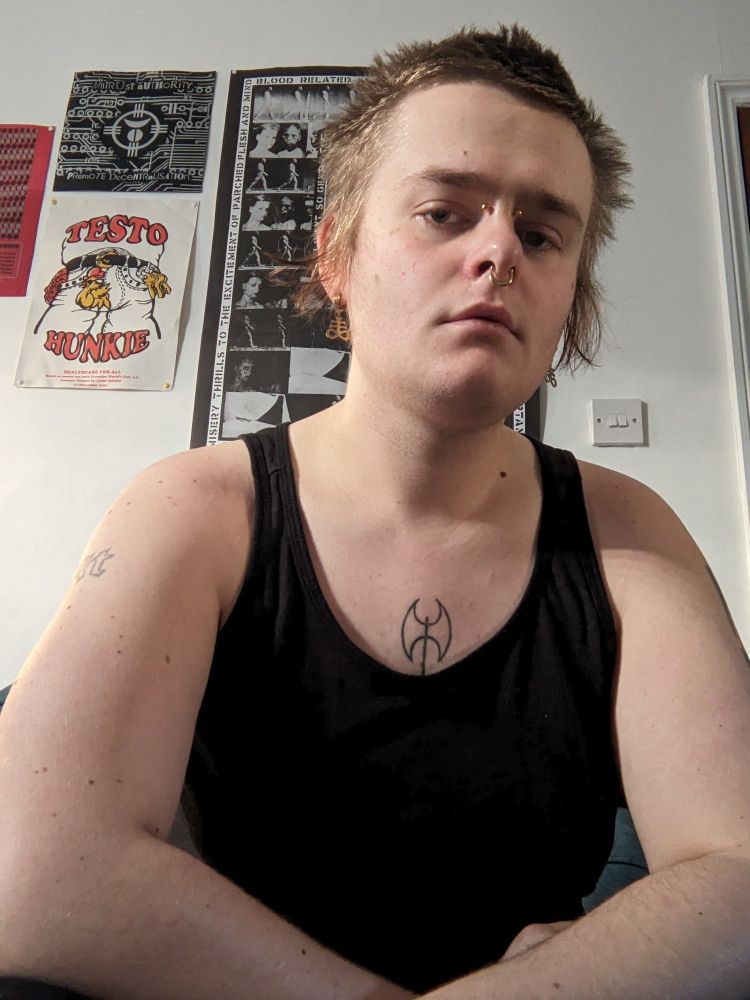 butch trans woman with boyish scrungly bed hair, gold facial piercings. wearing a black tank top which exposes a labrys chest tattoo.