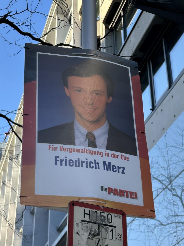 Das Bild zeigt ein politisches Plakat, das an einem Laternenmast befestigt ist. Es enthält ein Porträt eines Mannes im Anzug mit Krawatte. Auf dem Plakat steht der Satz: „Für Vergewaltigung in der Ehe“ gefolgt von dem Namen „Friedrich Merz“. Darunter befindet sich das Logo der Partei „Die PARTEI“.

Das Plakat scheint satirisch zu sein, da „Die PARTEI“ für satirische politische Botschaften bekannt ist. Im Hintergrund sieht man eine städtische Umgebung mit einem Gebäude und einem blauen Himmel.

