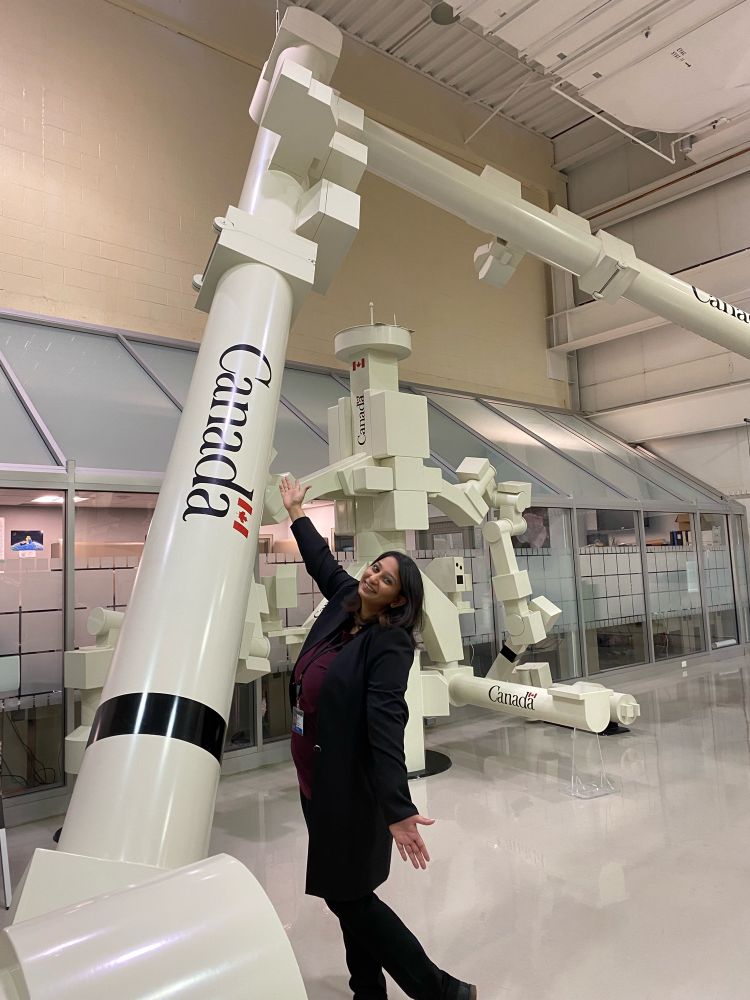 Parshati is smiling and standing with her arms open wide side ways in front of a life size mock up of Canadarm. 