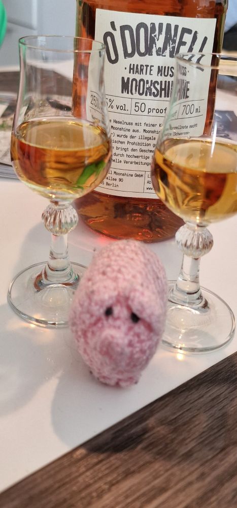 Ein süßes, rosa Schweinchen gehäkelt aus rosafarbener Wolle, steht zwischen 2 Schnapsgläsern und guckt niedlich in die Kamera