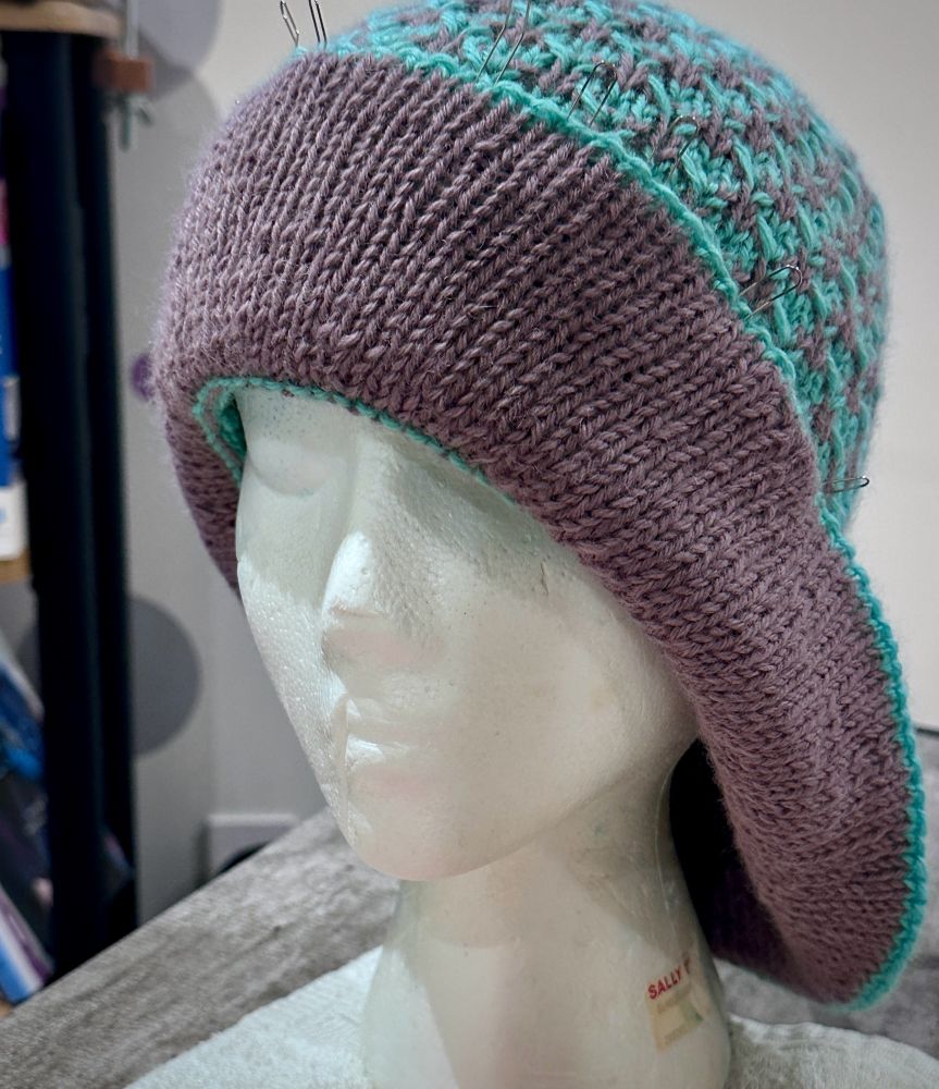 Hand knit cloche hat shaped on styrofoam mannequin. Cloche hat worked in texture two color slip stitch pattern of green and purple Hat brim partially turned up and pinned revealing purple side of brim. 