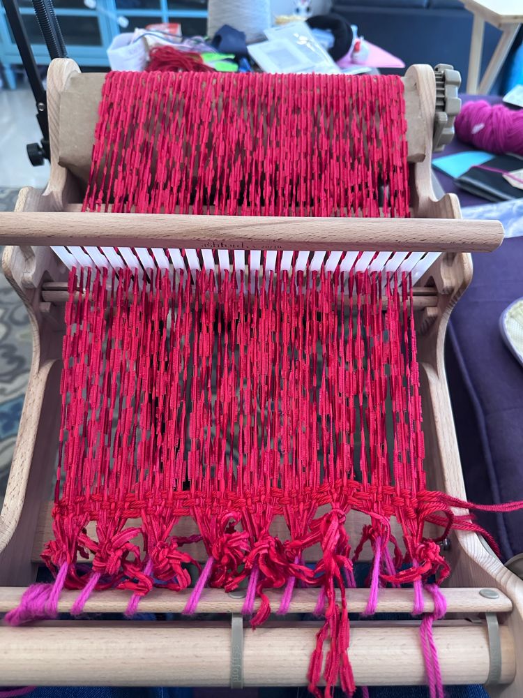 Rigid heddle loom warped and ready to weave. Warp is a red novelty yarn lashed onto front beam with pink yarn. 