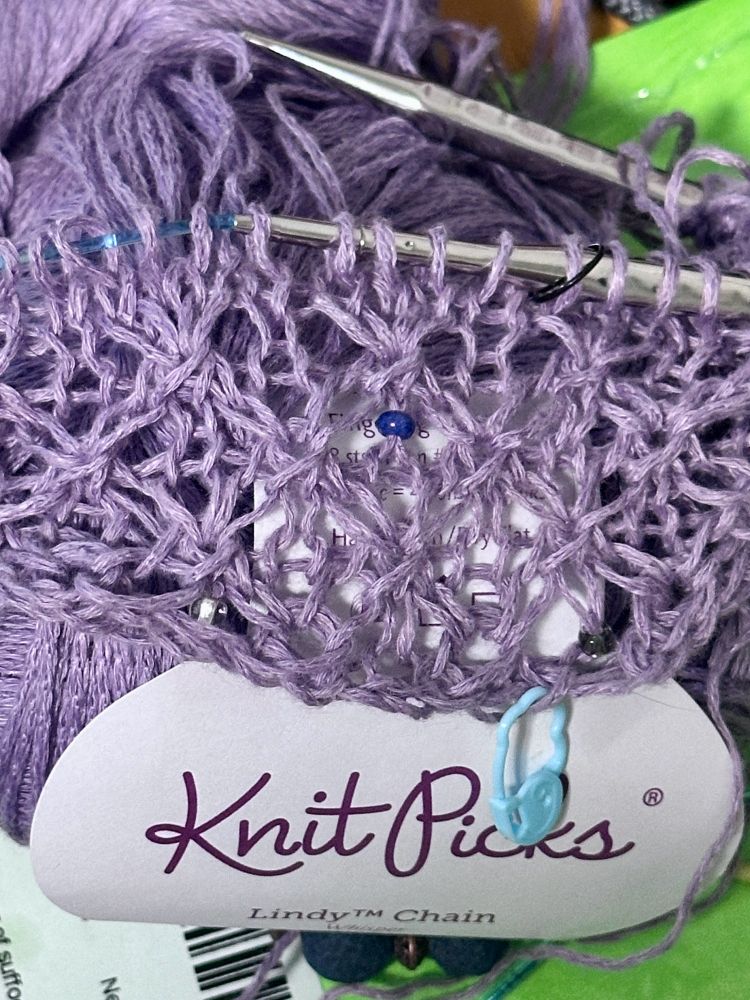 Work in progress of hand knit lacy shawl worked in lilac yarn and featuring a purple bead sits on ball of lilac color yarn.