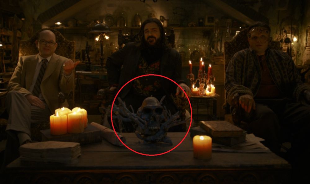 A screenshot form the series What We Do In The Shadows depicting two vampires and a Frankenstein-esque monster seated in three chairs talking directly to camera in a spooky candle-lit vampire house. On the table in front of them, centred in the shot and surrounded by old books and candles, is a cast of a skull of Australopithecus afarensis, situated in a small dead tree or something like a table centrepiece.