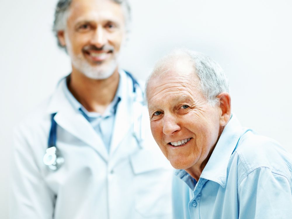 An older patient smiling at the camera with a healthcare professional standing behind them, both in a clinical setting.
