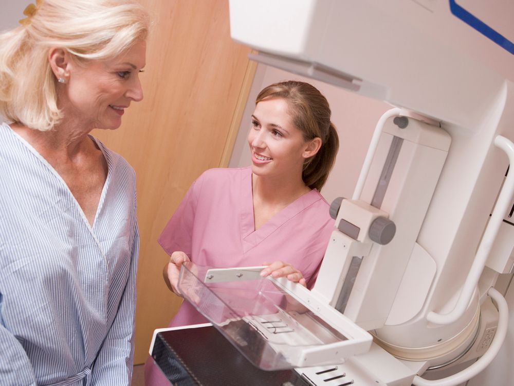 A healthcare professional wearing pink scrubs talking with a patient in a light blue gown at a mammography machine.