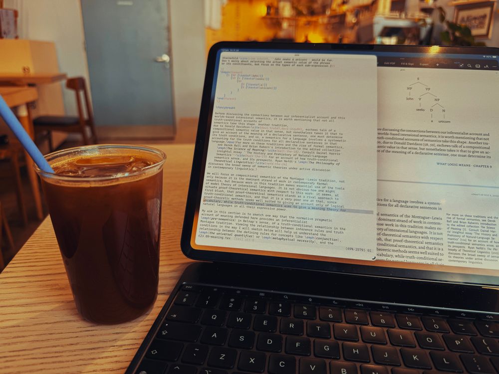 A clear glass filled with cold black coffee, ice, and containing a green straw, to the right of an iPad, with two windows open, one a Vim session, editing a latex file, and the other, the pdf of the compiled output. A parse tree of the sentence “John seeks a unicorn” is visible in both views.