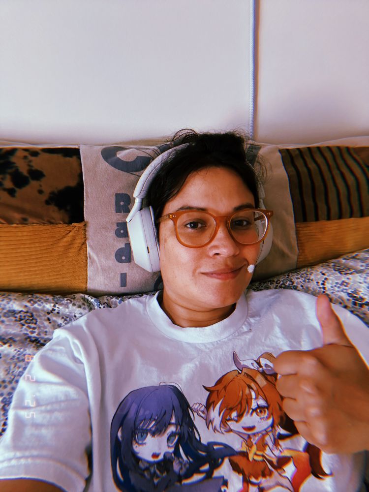 this is me, hana, giving a thumbs up in front of my cool new pillow! i have my headphones on and a daisy-shaped acne patch covering a ~blemish~ on my left cheek. also my shirt has crunchyroll hime and ado on it which is irrelevant to anything. :3