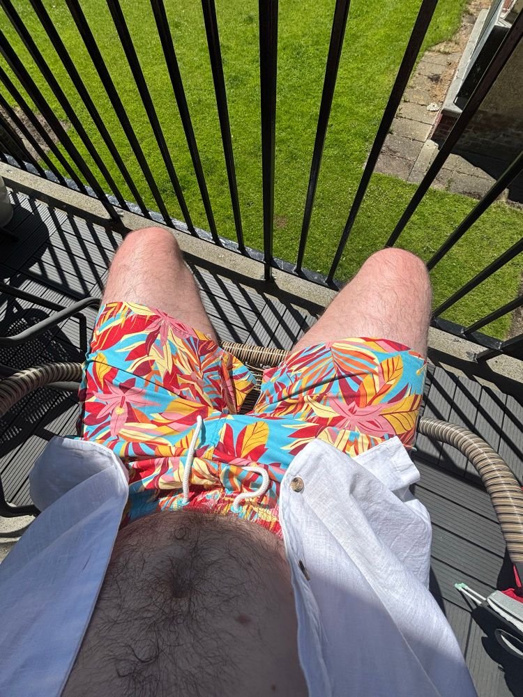 My camera is looking down from a balcony as vibrant green grass, focus is on my fun swim shorts glistening in the sunshine on the balcony. Shorts have a red and yellow leaves design against a light blue colour 