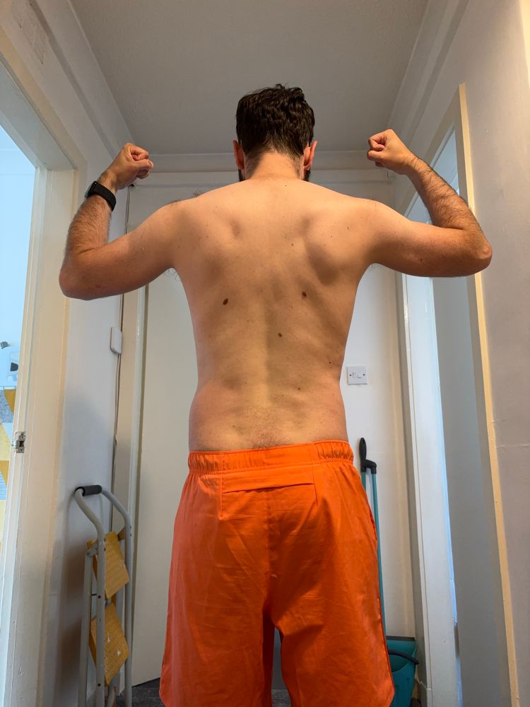 A rear camera photo of my back and gym progress, my arms are tensed up in the air and I have my coral gym shorts on