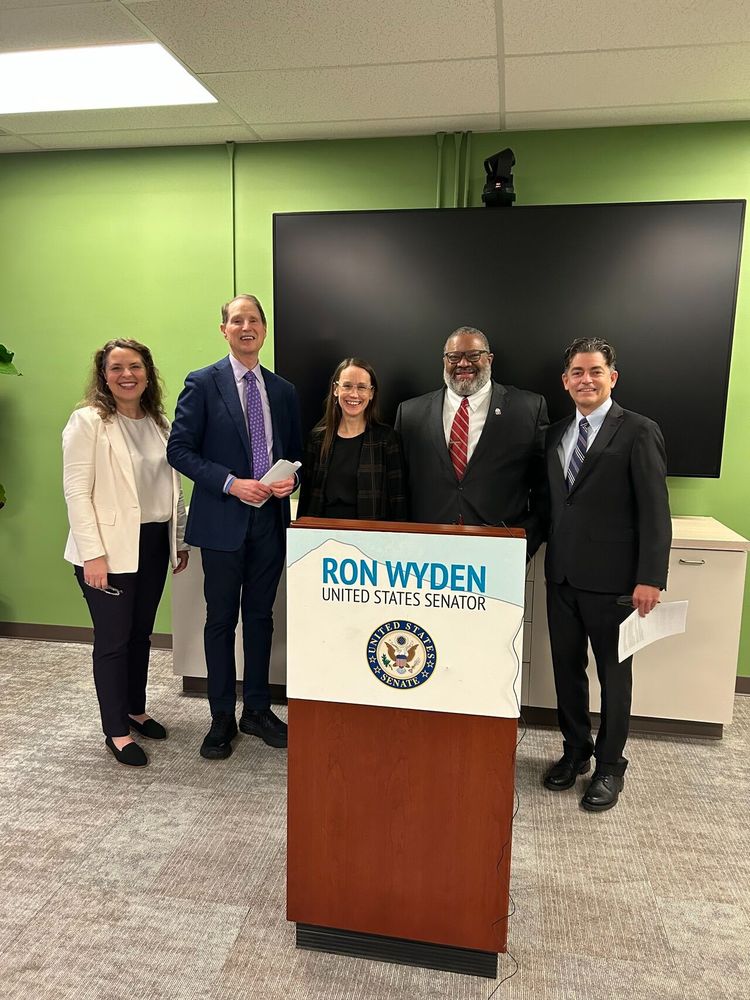 Against a green background and TV screen, Senator Wyden and smiling group take photo after press conference.