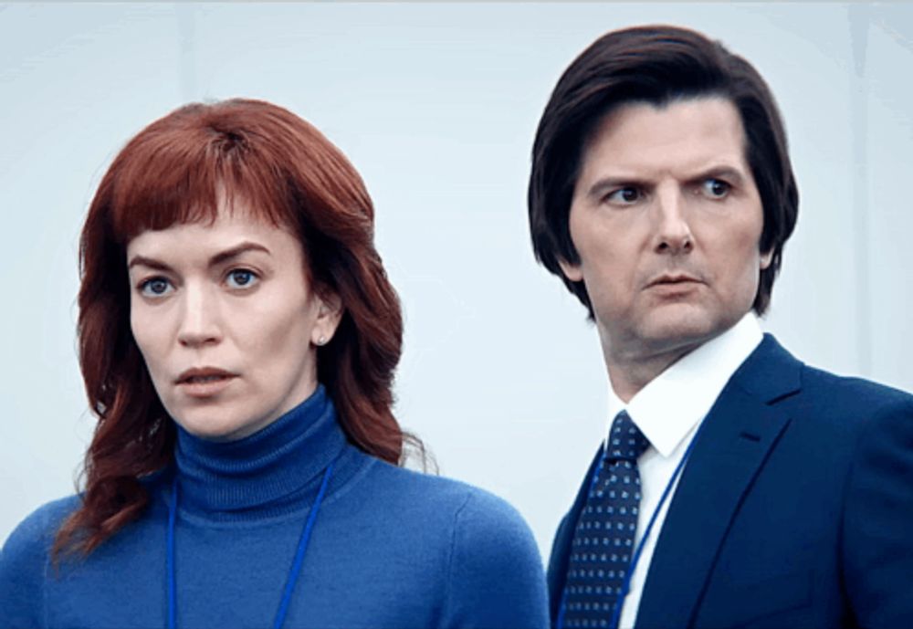 a man in a suit and tie stands next to a woman in a blue turtleneck