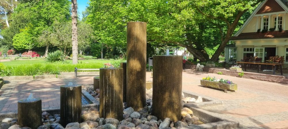 Ein Springbrunnen im Stadtpark von Nordhorn. Fünf unterschiedlich hohe Säulen, aus denen oben Wasser sprudelt. Rechts ein altes Haus, im Hintergrund die sehr schönen Bäume.