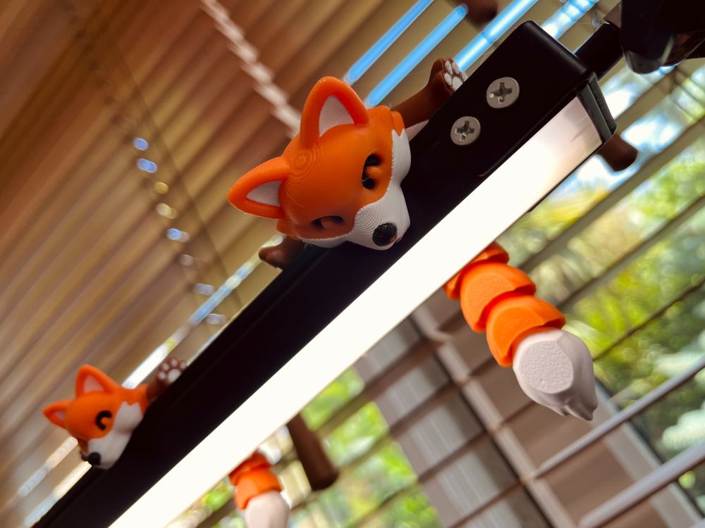 A pair of orange and white plastic foxes with articulated tails sitting on top of a flat desk lamp, in front of a blind-covered window through which we can see an out-of-focus sunny garden scene.