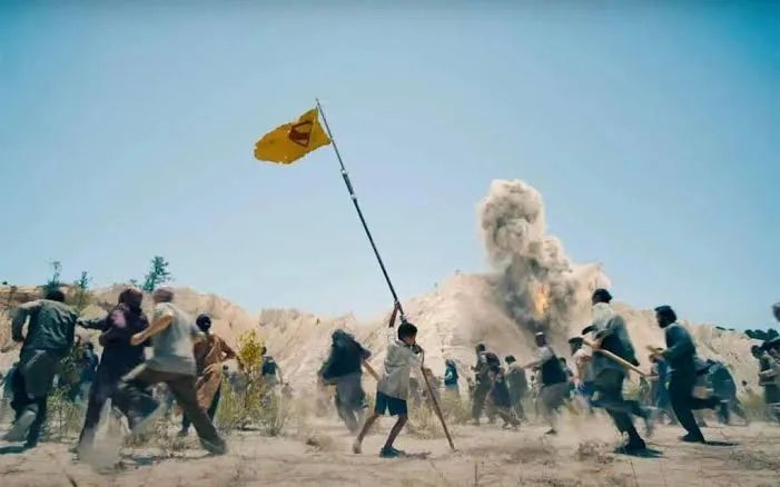 Image from Superman 2025. As people flee all around, bombs and shells dropping, a little brown boy holds up a home made flag of a yellow background with a red S for Superman. 