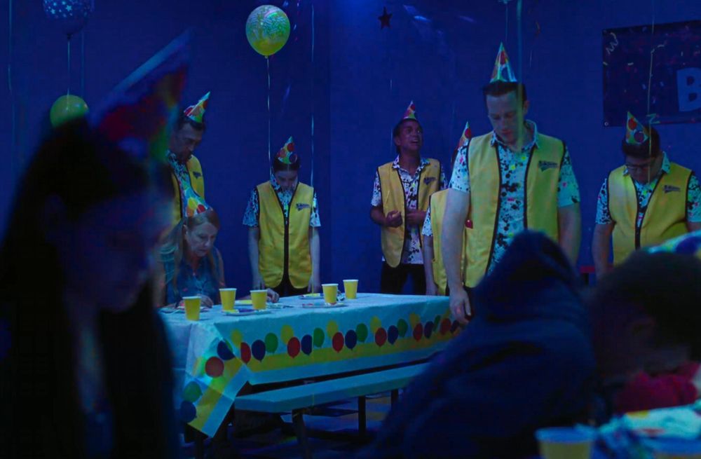 A scene of a children's birthday party at a family fun center, crowded with employees whose heads are all slumped as they stand, frozen. In the back, a man looks desperately to the ceiling.

