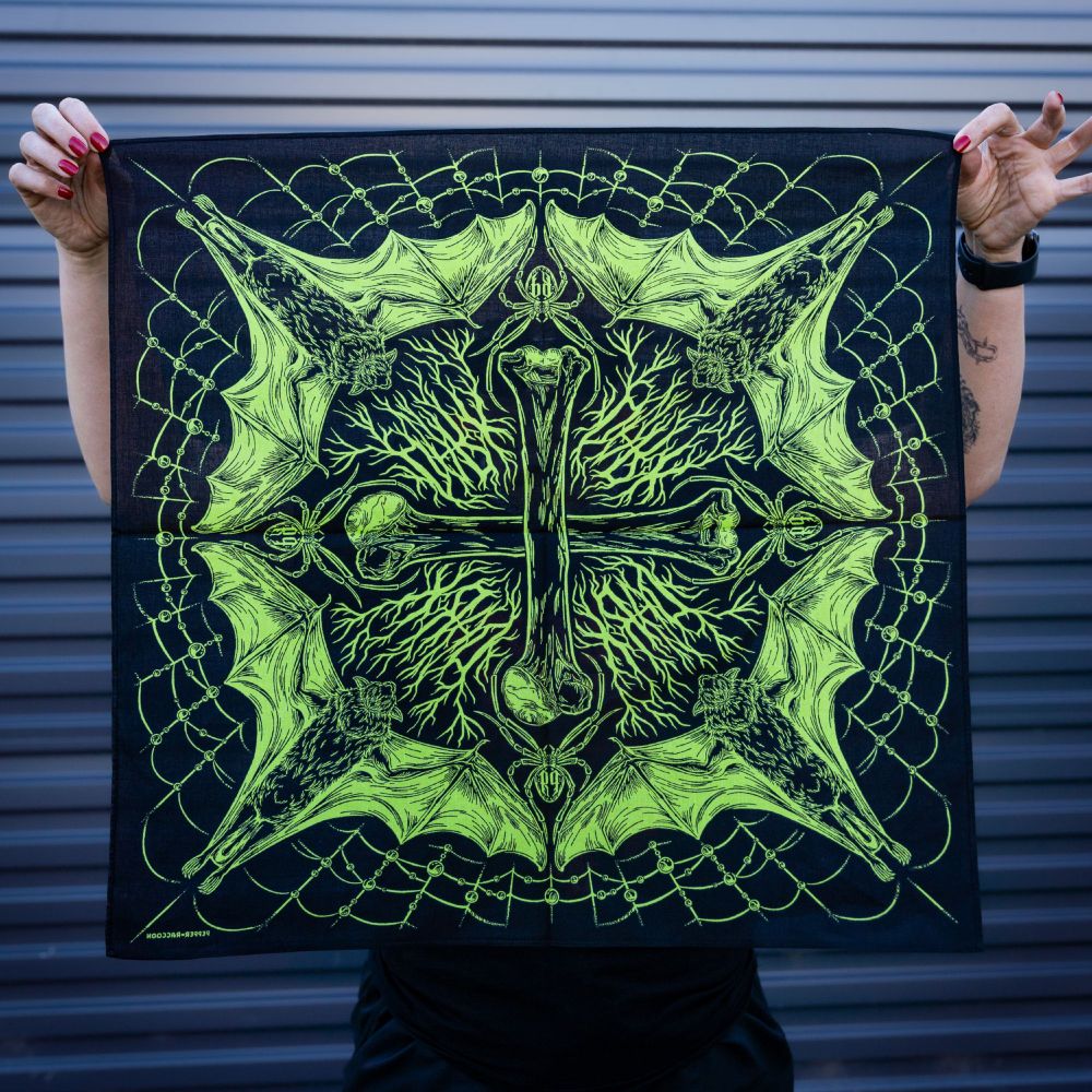 A person holding up a black bandana with lime green spooky illustrations of bones, bats, and spiderwebs