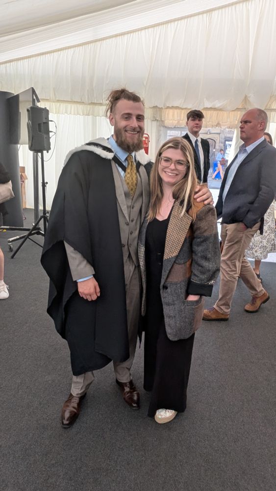 Two smiling siblings in a marquee, the brother is wearing graduation robes 