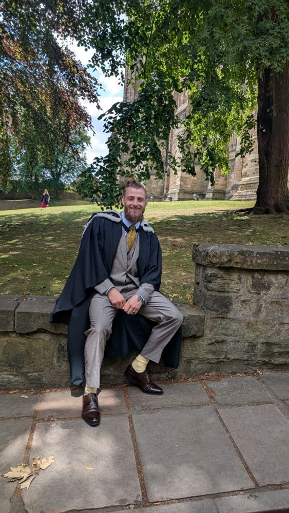 A young man in Durham University graduation robes sitting on a wall and smiling 