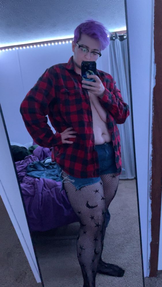 Indrid stands in front of a mirror taking a selfie. They are wearing fishnet tights with stars and moons in the pattern, denim short shorts with the side hems cut open and corseted with chains, and a red and black buffalo check flannel shirt. 