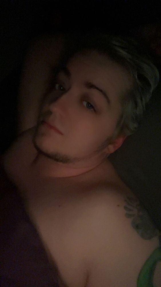 Indrid is shirtless in bed with a purple blanket pulled up to cover most of their chest. Parts of their tattoos on their right arm are visible. The room is dark and they look sleepy and comfortable